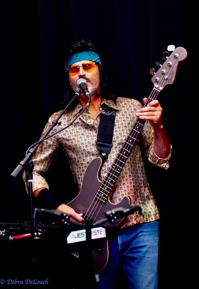 Musician singing into a mic while playing a purple bass guitar on stage, wearing a patterned shirt and blue jeans