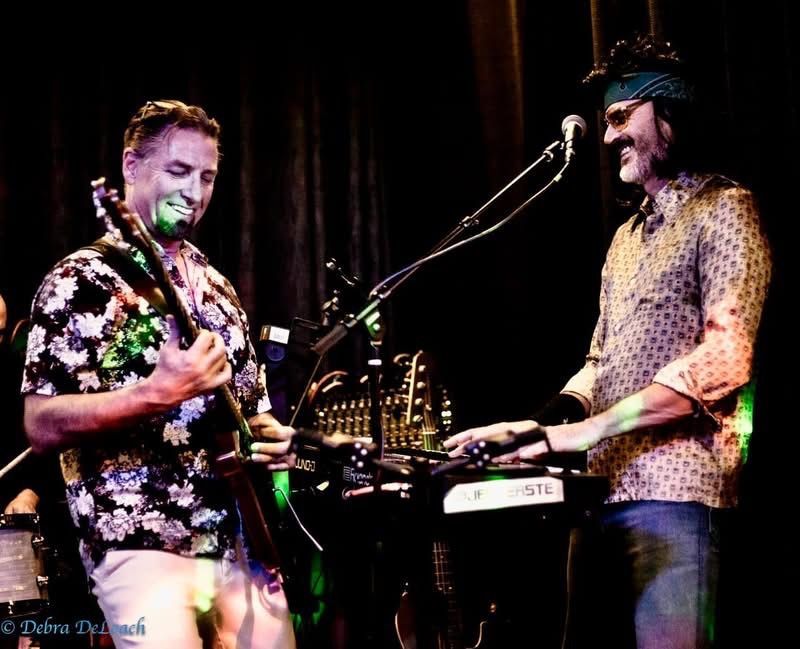 Two musicians performing onstage, one playing guitar and one at a keyboard under colorful lights.