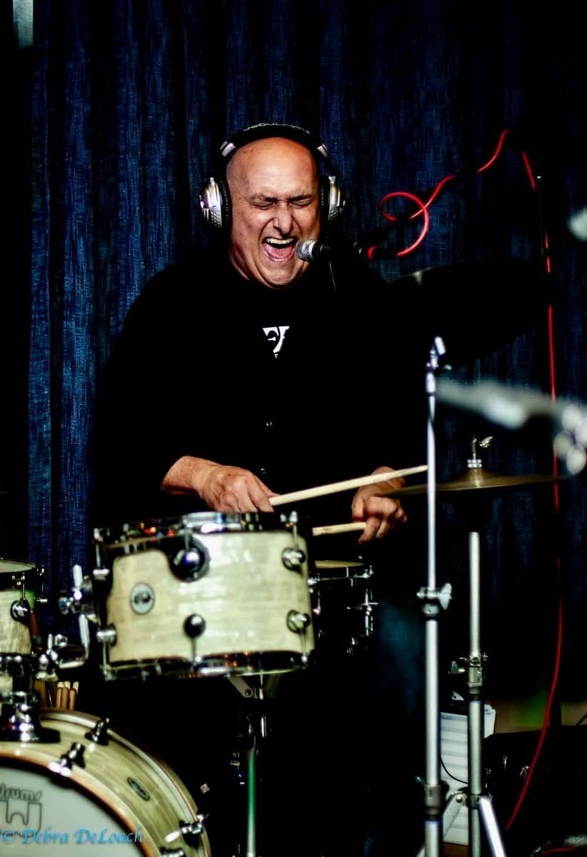 Drummer wearing headphones plays a cream drum kit on a dark stage, smiling under studio lights