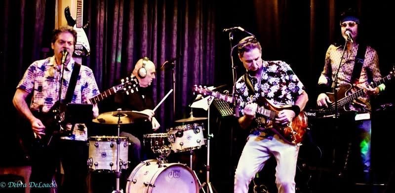 Four musicians perform onstage under purple lighting, with guitars, drums, and a microphone.