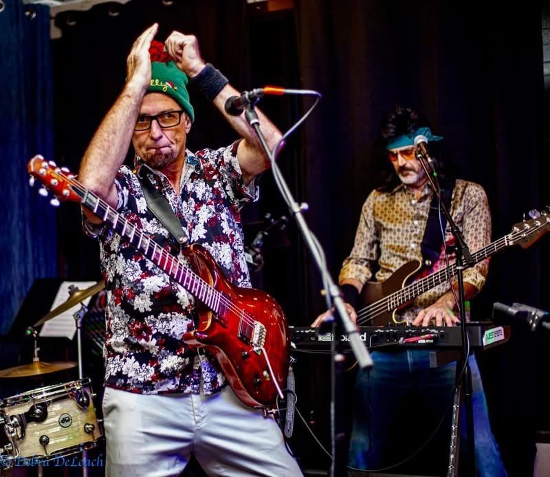 Two musicians performing onstage, one raising his hands, with red and black electric guitars and microphone stands.