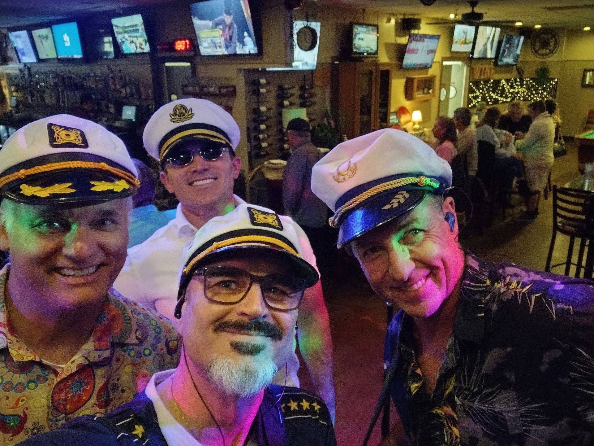 Four smiling people in captain hats pose in a lively bar with colorful lights and TVs in the background