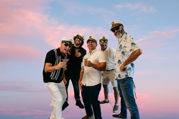 Five friends posing together on a beach at sunset, with pink and blue sky behind them.