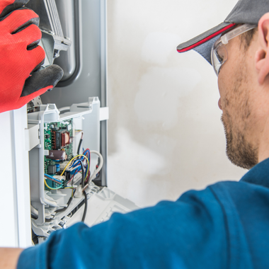 Technician in blue shirt, safety glasses and cap, repairs a circuit board inside a white appliance, red gloved hand present.