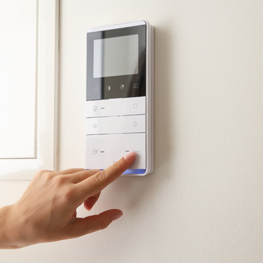 Hand pressing a button on a white intercom panel mounted on a wall.