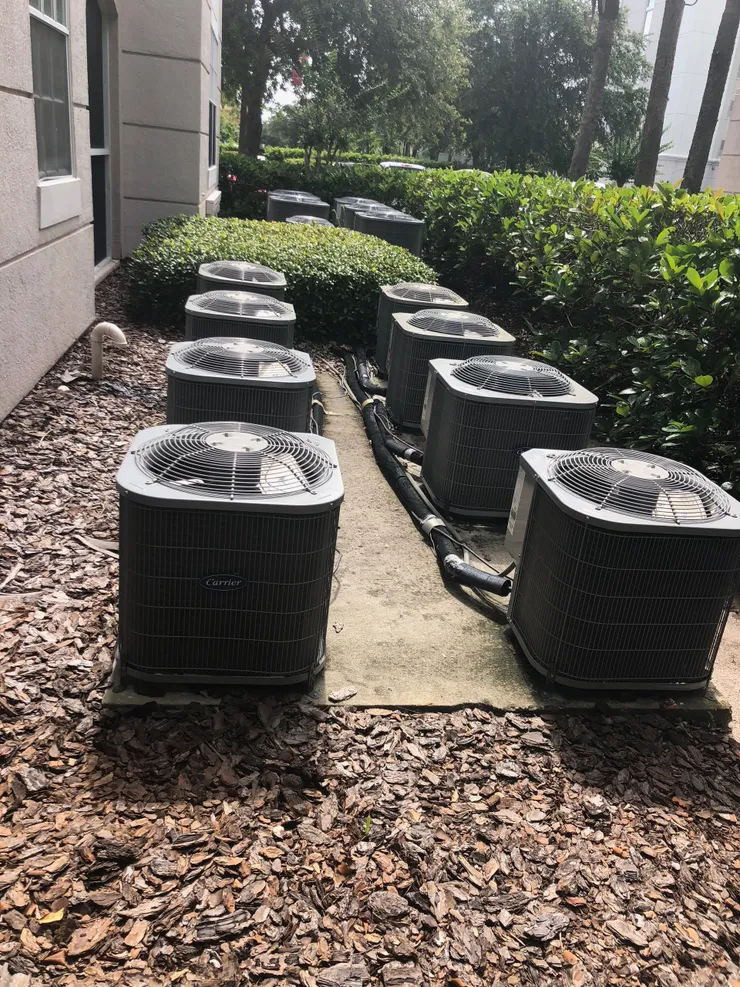 Row of outdoor air conditioning units near building and foliage.