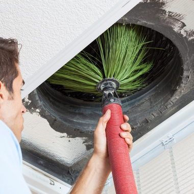 Person cleaning air duct with a brush tool. The duct is open in the ceiling.