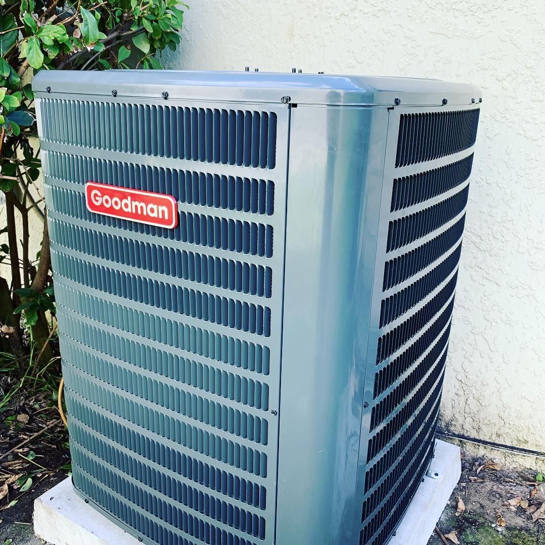 Goodman air conditioning unit outside on a concrete base, with a white wall and green foliage in the background.