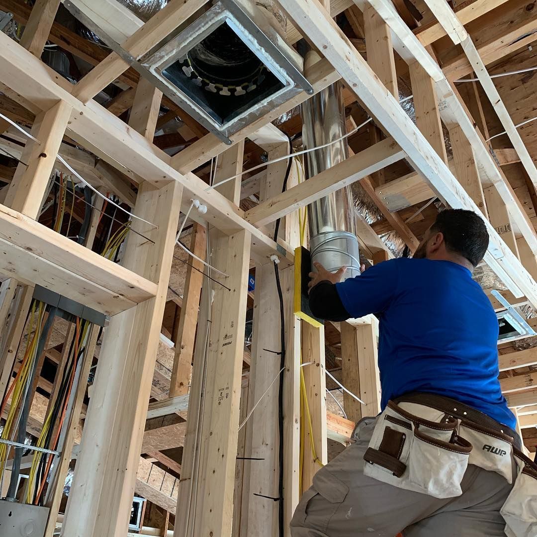 Construction worker installing ductwork in a wood-framed ceiling. He's wearing a blue shirt and tool belt.
