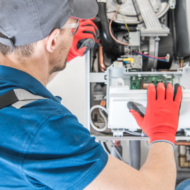 HVAC technician working on a furnace, wearing red gloves, cap, and glasses.