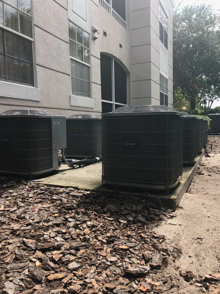 Air conditioning units lined up outside a building with windows, set on concrete pads.
