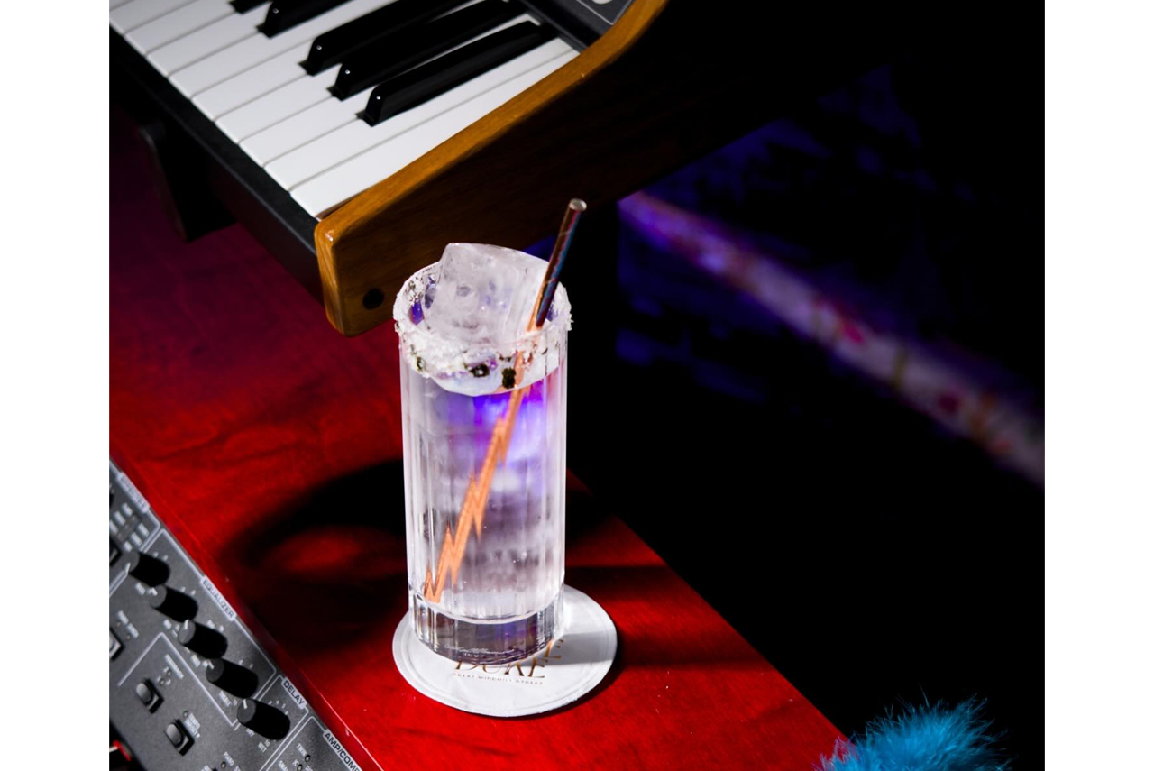 Glass of iced cocktail with straw on a red table beside a keyboard, under purple lighting