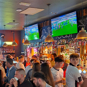 Crowded pub with patrons watching soccer on large TV screens under warm hanging lights