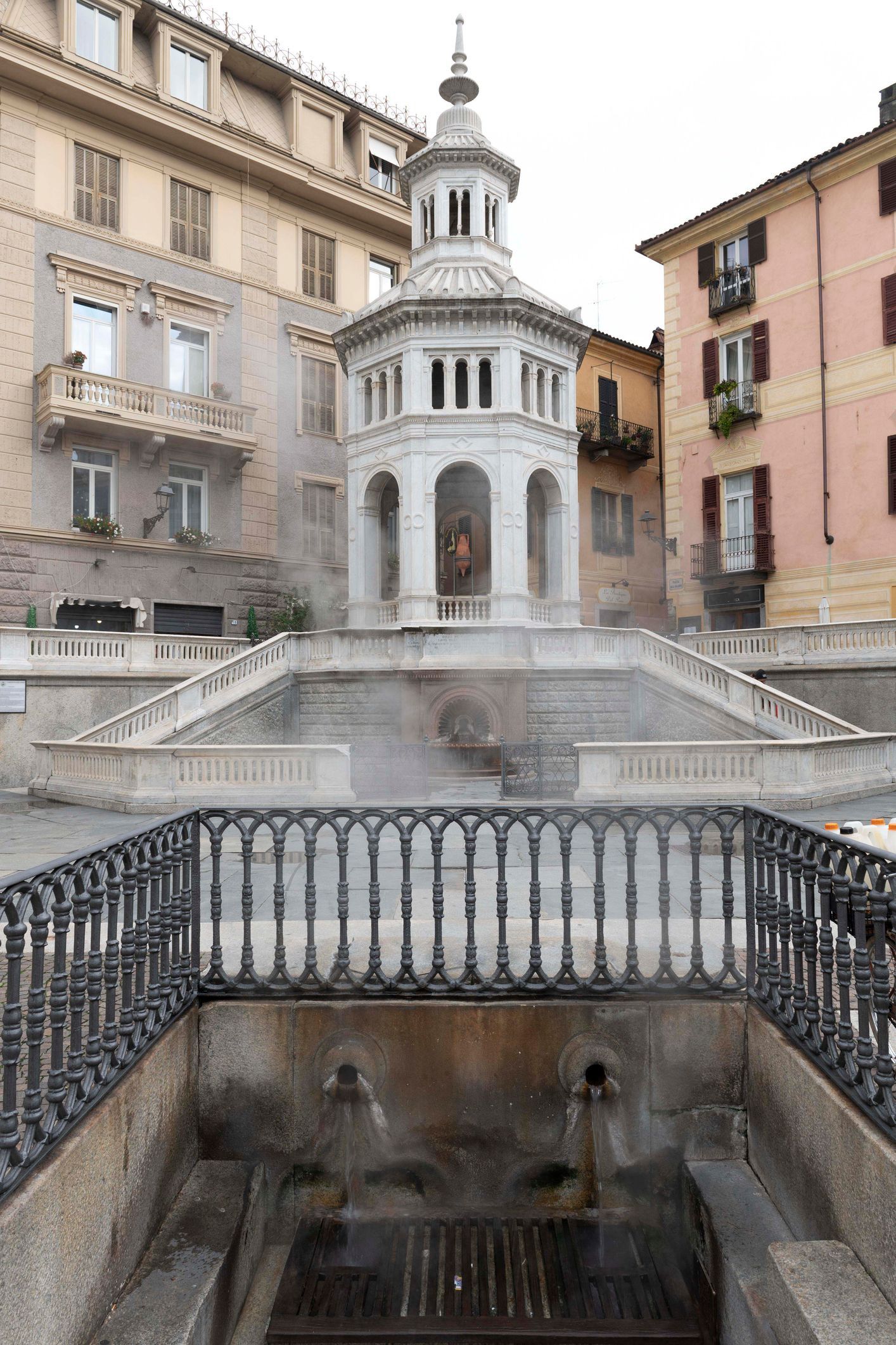Elegante fontana in pietra bianca in una piazza europea, incorniciata da edifici storici e ringhiere in ferro.