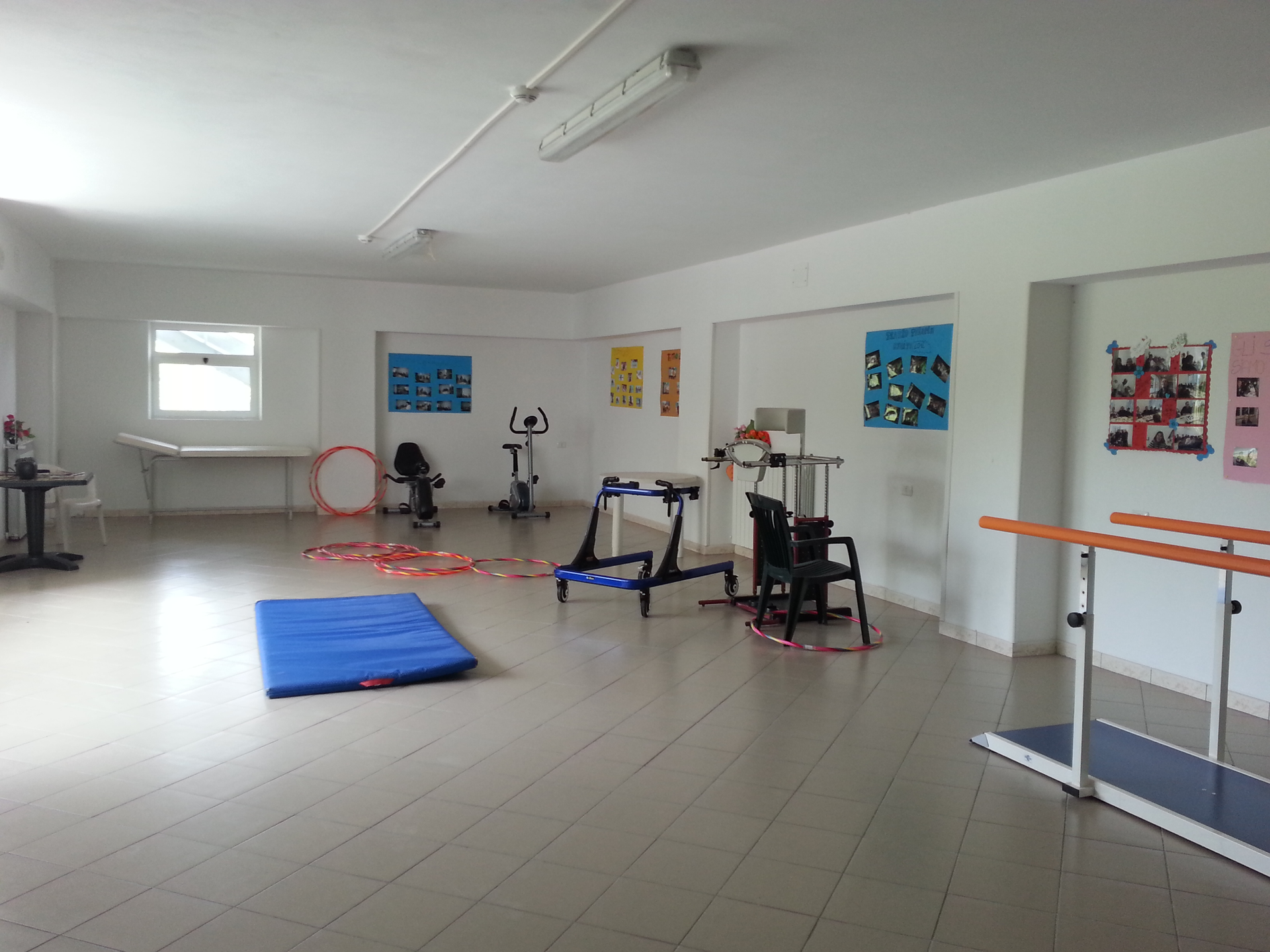 Sala di fisioterapia vuota con attrezzi per esercizi, tappetini e parallele su un pavimento piastrellato.