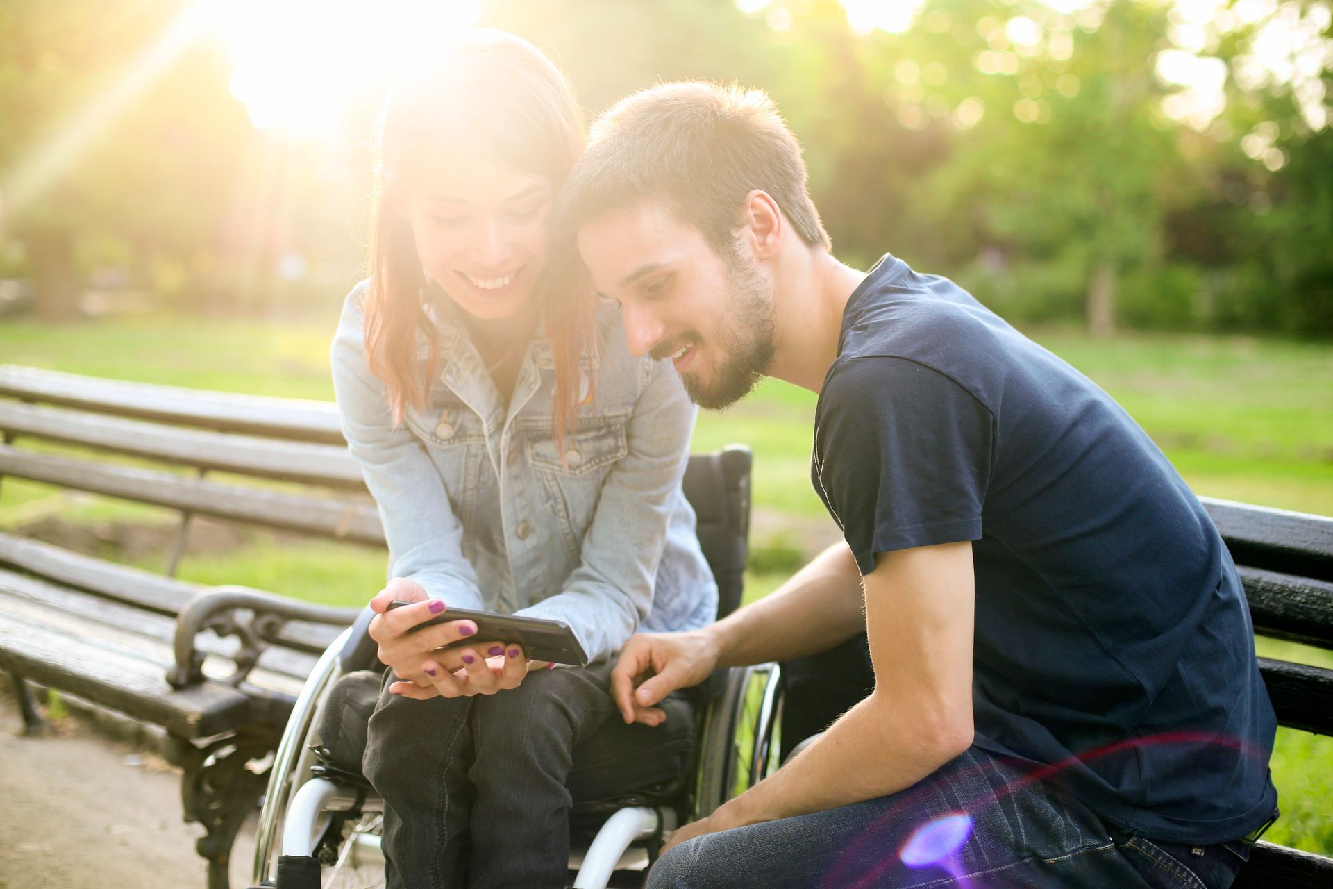 Due persone all'aperto in una giornata di sole, sorridenti sedute accanto a una sedia a rotelle in un parco.