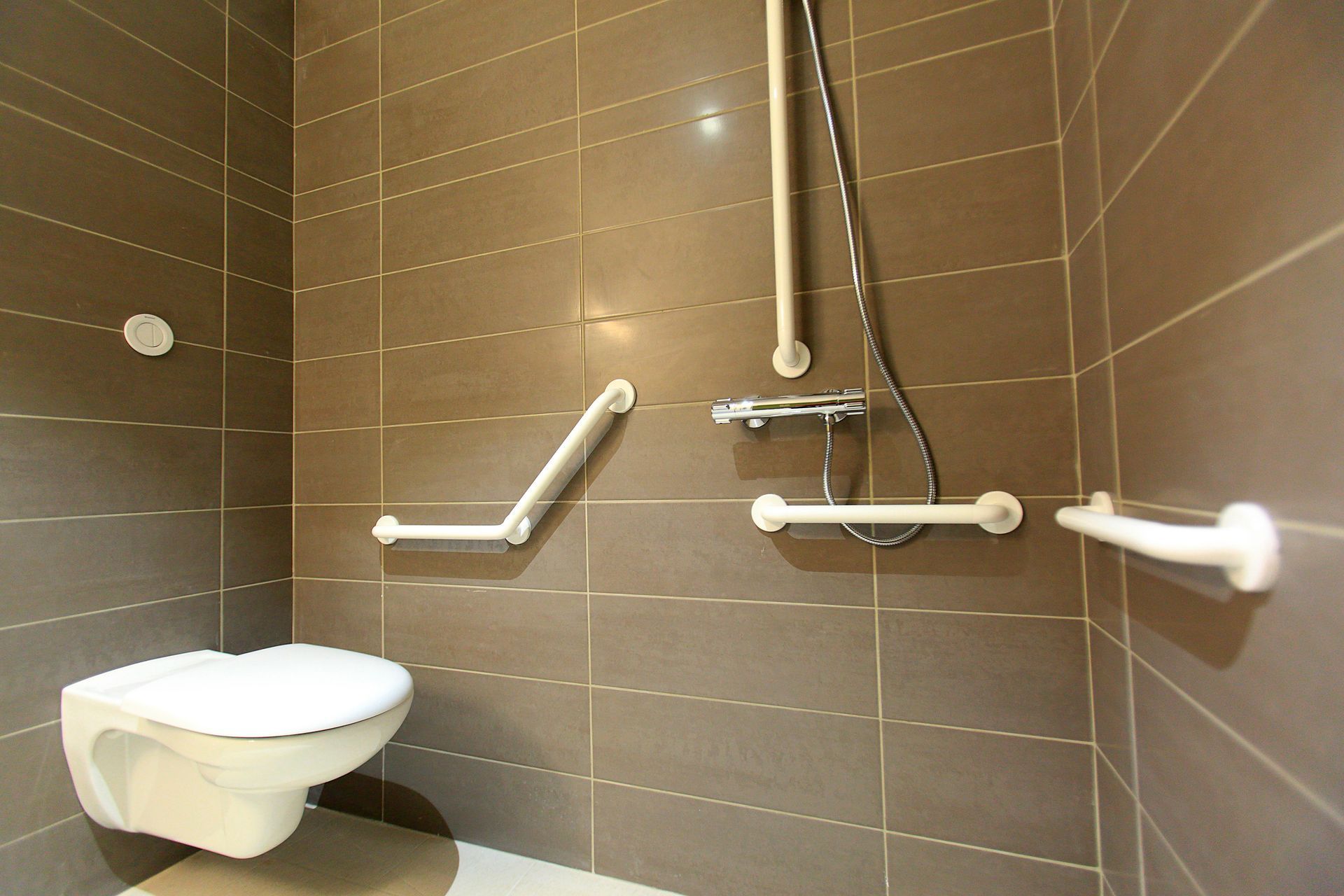 Bagno accessibile con WC a parete e maniglioni di sostegno in acciaio inox su pareti rivestite di piastrelle marroni.