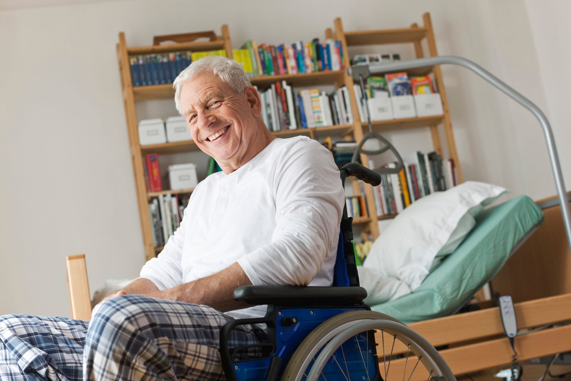 Un uomo sorridente in sedia a rotelle accanto a un letto d'ospedale in una stanza con scaffali pieni di libri.