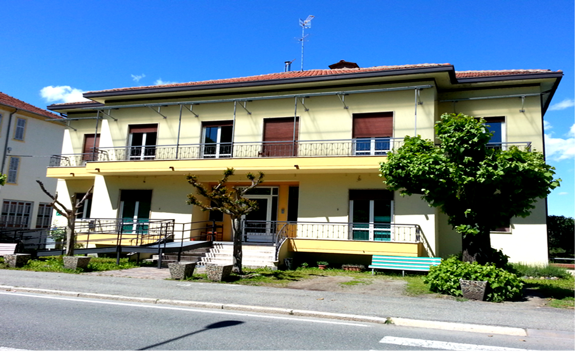 Casa gialla a due piani con balconi e un albero a bordo strada sotto un cielo azzurro limpido
