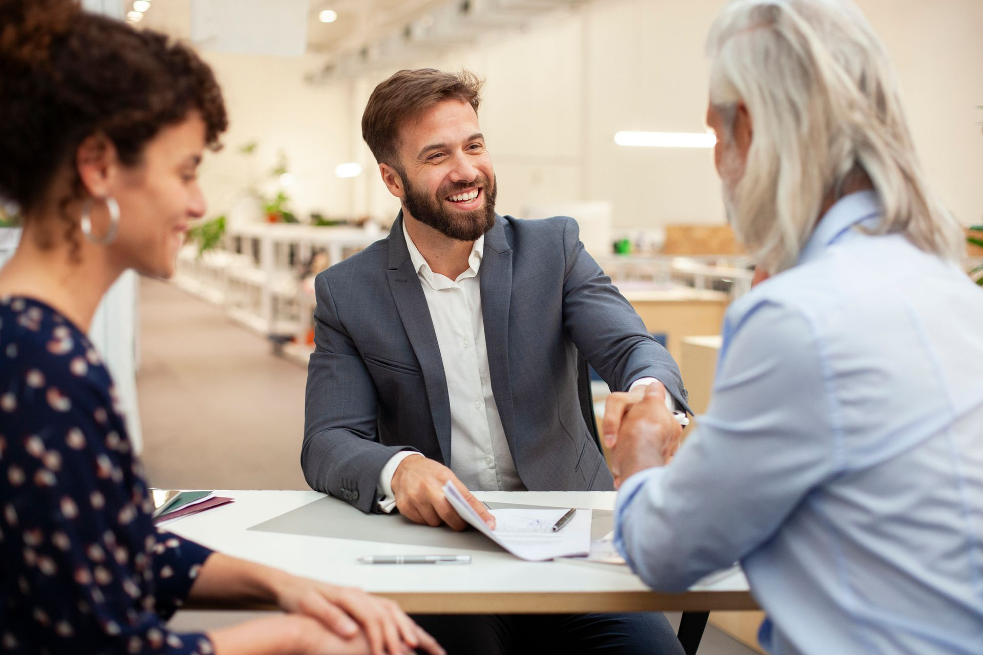 Tre persone, sorridenti e concentrate, discutono di documenti attorno a un tavolo in un ufficio luminoso.