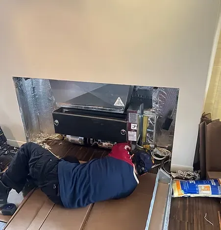 A person in a blue shirt works on a built-in appliance in a wall, with insulation visible.