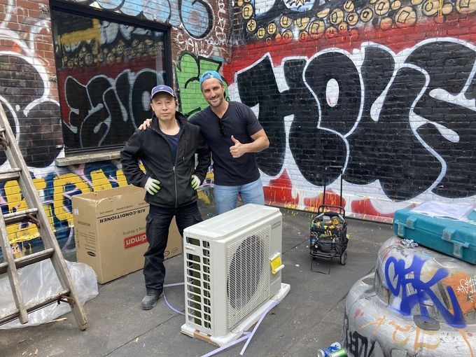 Two men stand with an air conditioner in a graffiti-covered alley. One gives a thumbs up.