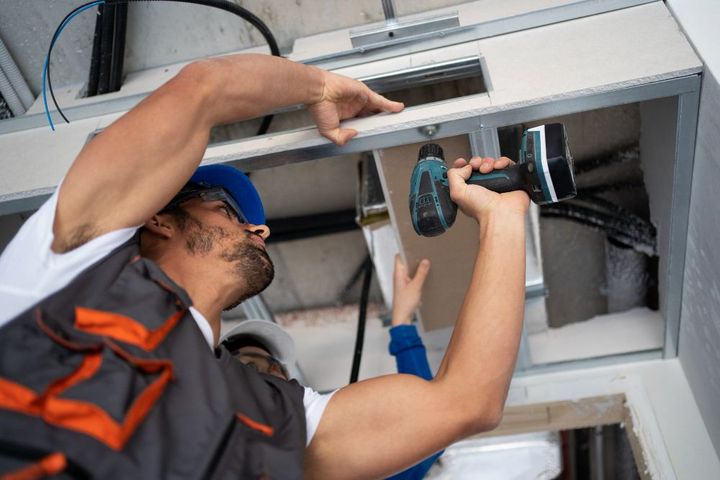 Construction worker installing drywall on a ceiling with a drill.