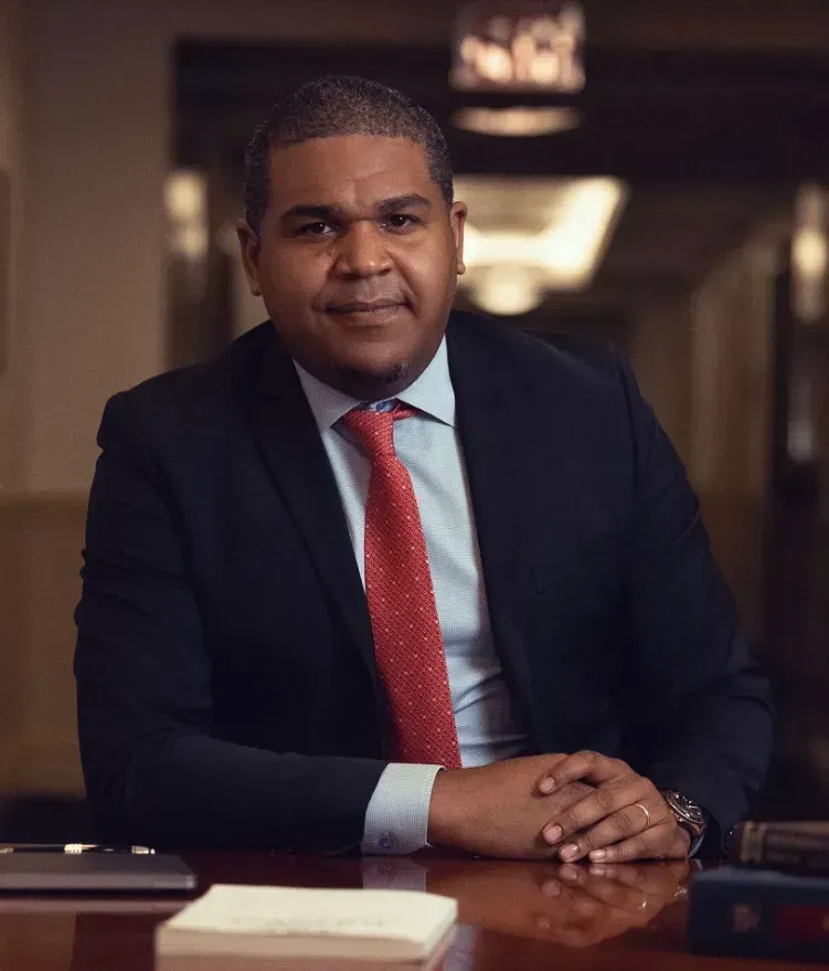 Man in a dark suit and red tie seated at a desk, hands clasped, in a softly lit office.