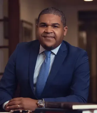 Man in a blue suit seated at a desk, smiling with hands folded in a warmly lit office.