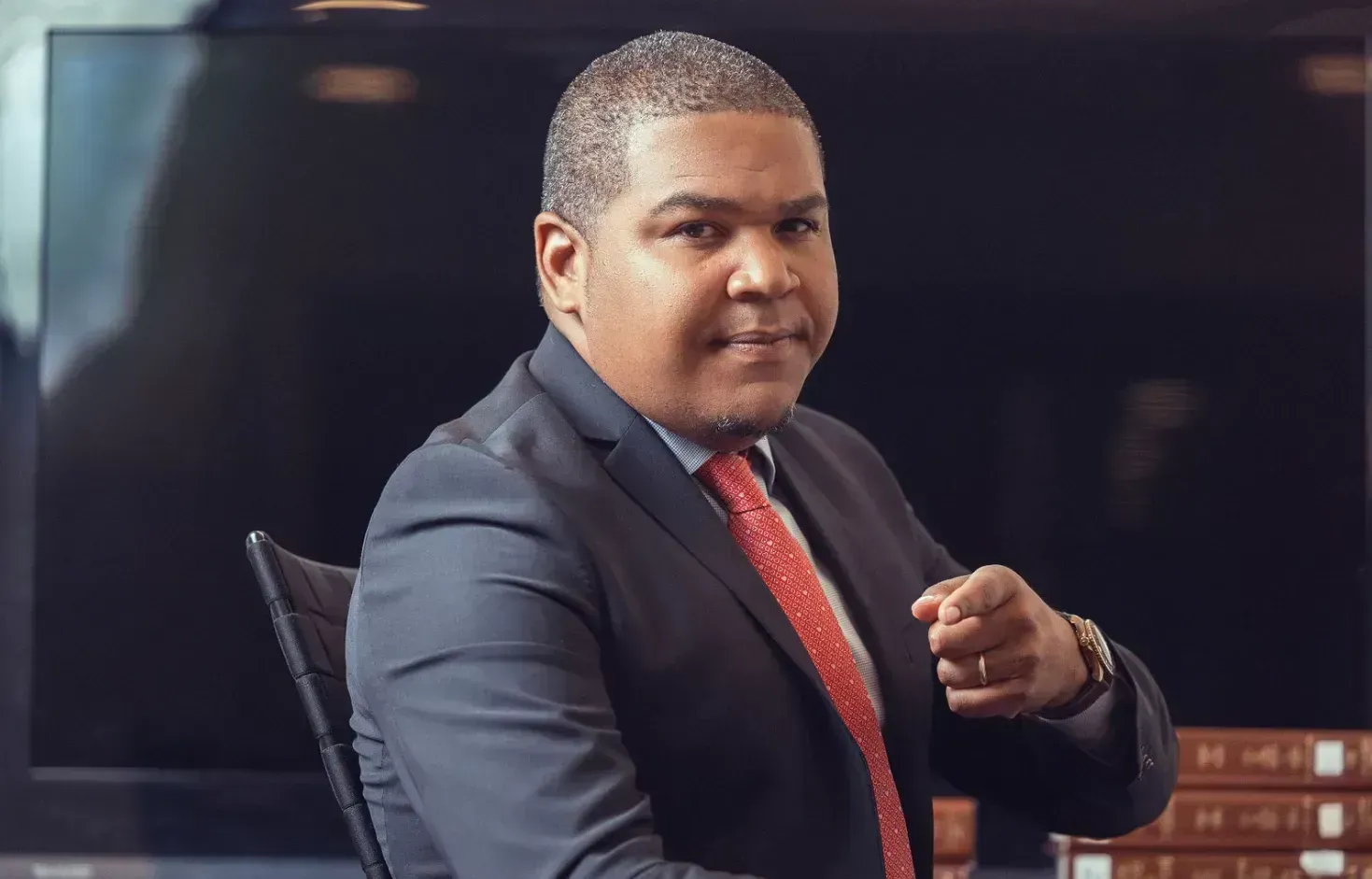 Man in dark suit and red tie seated at a table, looking at the camera with a raised fist.