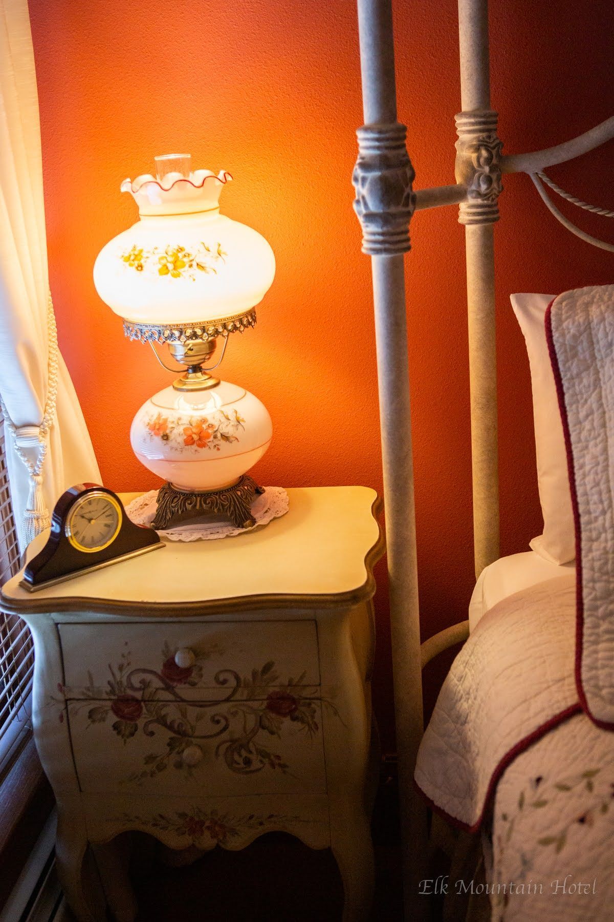 A lamp and clock are on a nightstand next to a bed.
