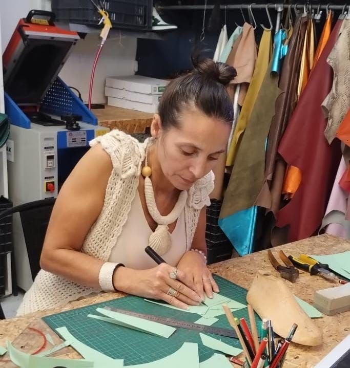 Mujer dibujando un patrón en papel verde en un banco de trabajo, con cuero y herramientas visibles.