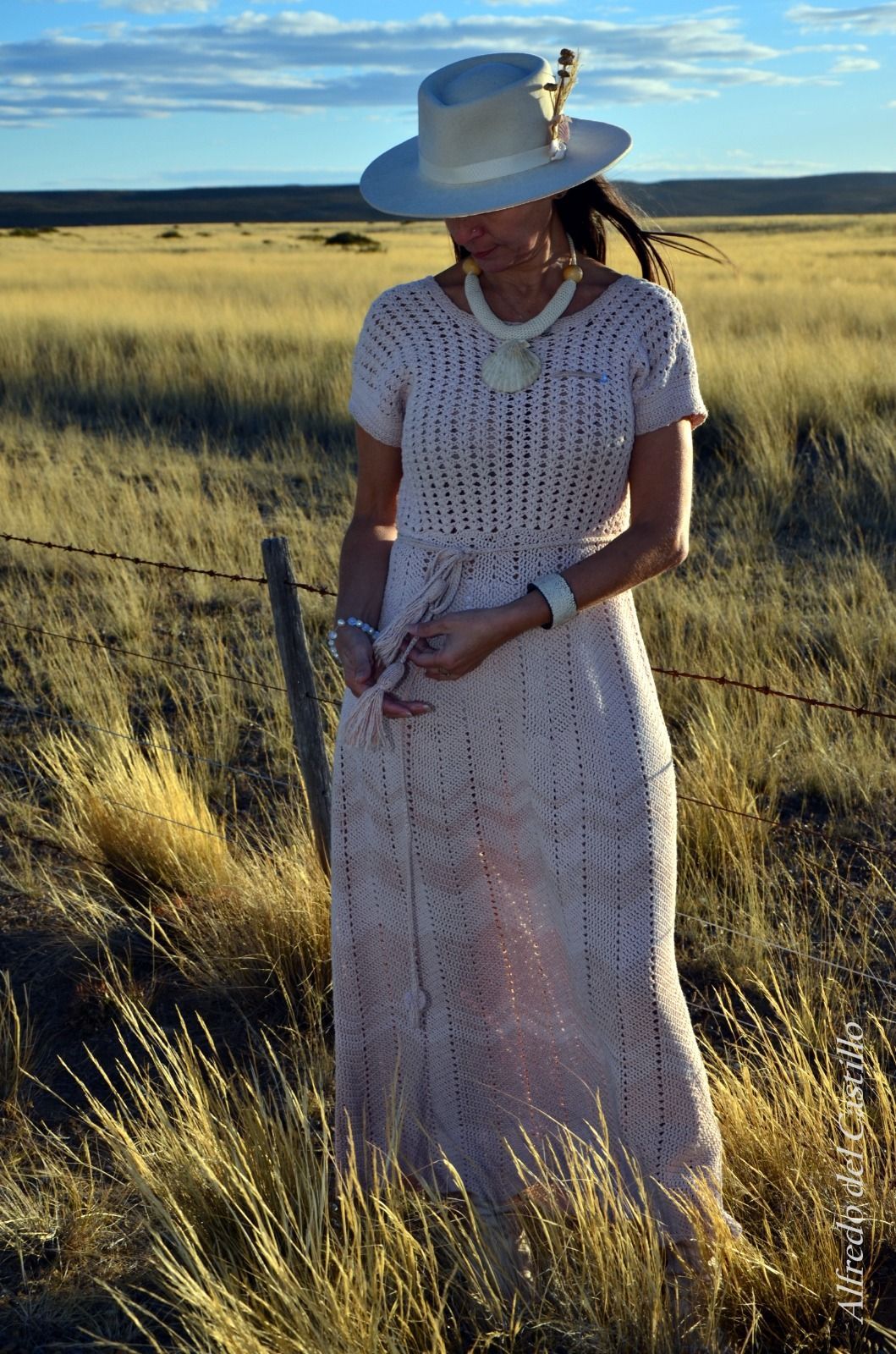 Una mujer con un vestido largo de color rosa claro y un sombrero está parada en un campo de hierba, cerca de una valla.