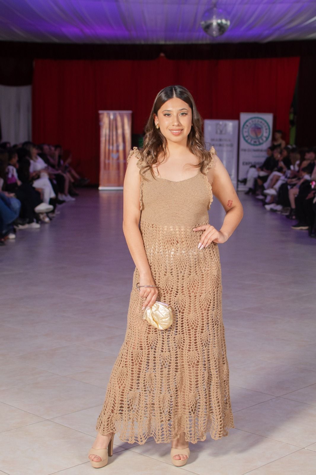 Mujer con un vestido de crochet beige en una pasarela, sosteniendo un clutch dorado.
