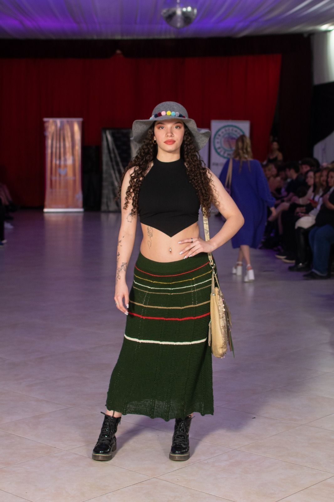 Mujer modelando una falda verde y un top negro en una pasarela, luciendo un sombrero, botas y bolso.
