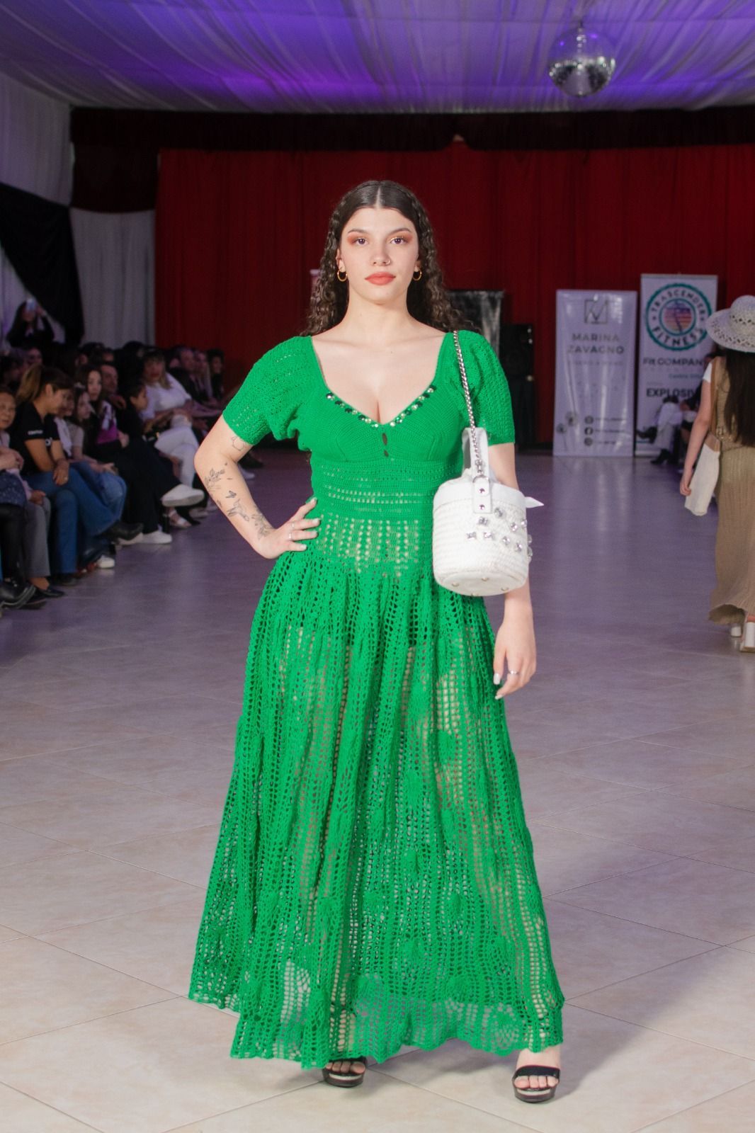 Mujer con un vestido verde y un bolso blanco, posando en una pasarela.
