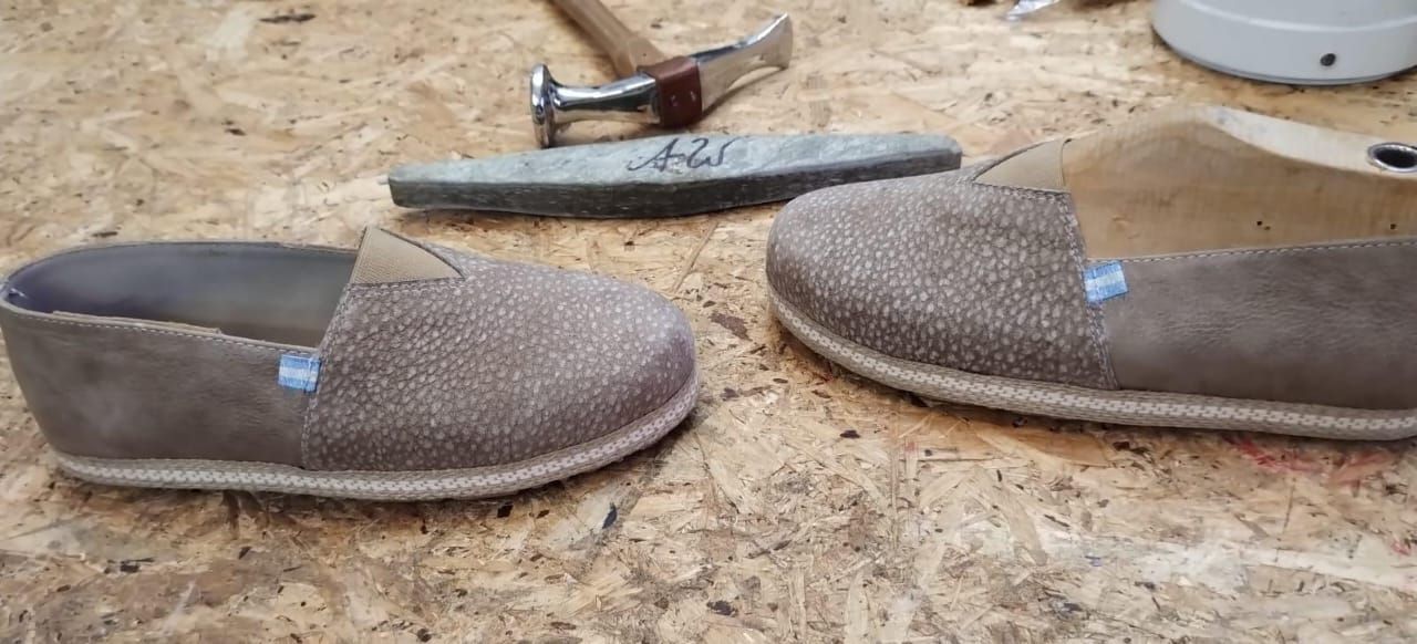 Dos zapatos marrones sin cordones con una herramienta al fondo. Los zapatos están sobre una superficie de madera.