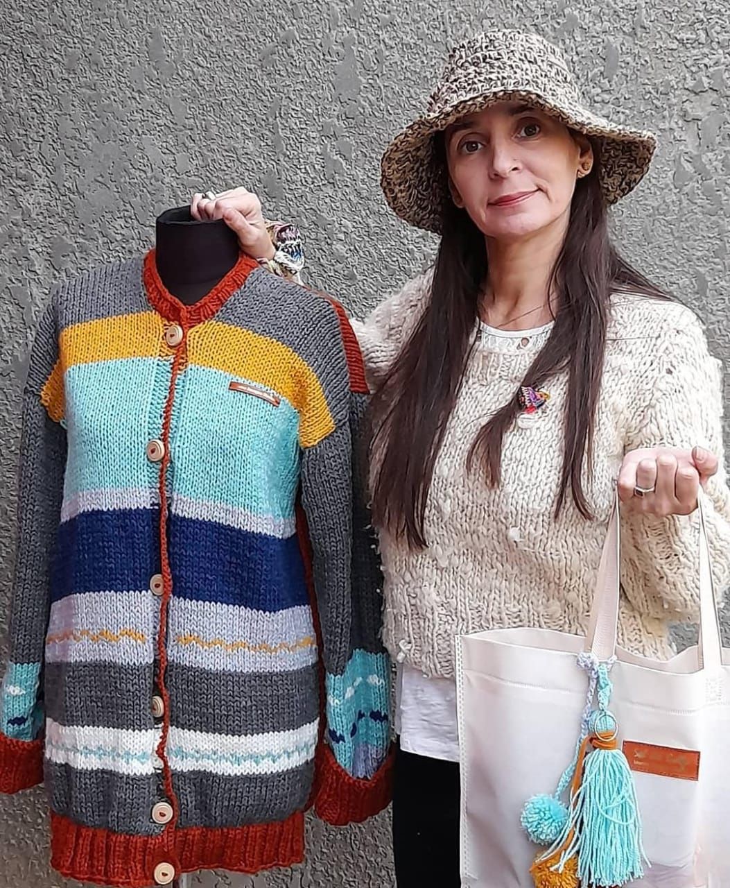 Mujer con cárdigan y sombrero hechos a mano, sosteniendo un bolso de mano, junto a un suéter a juego en un maniquí.