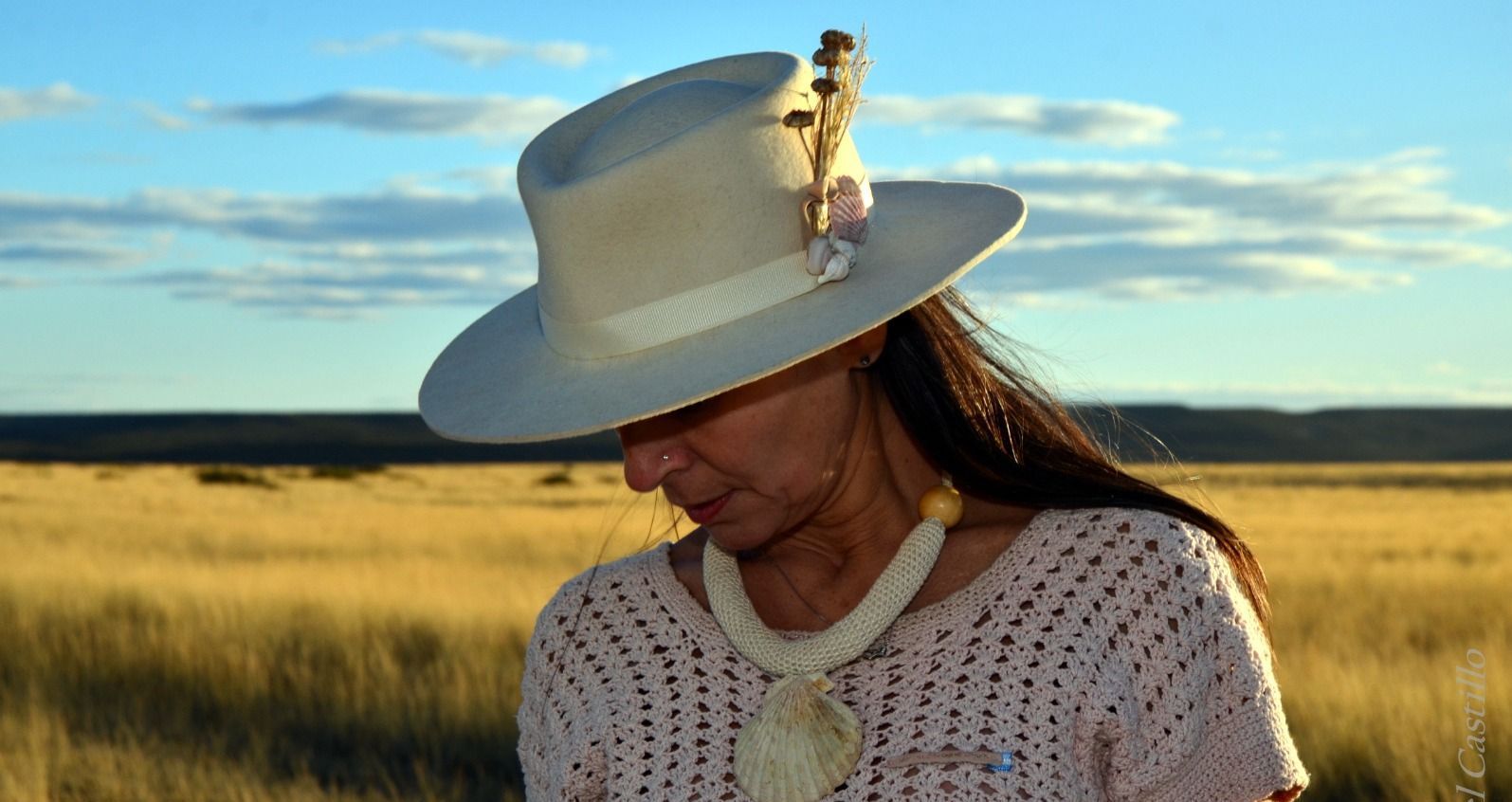 Mujer con un sombrero color crema, mirando hacia abajo en un campo de hierba seca, contra un cielo de atardecer.