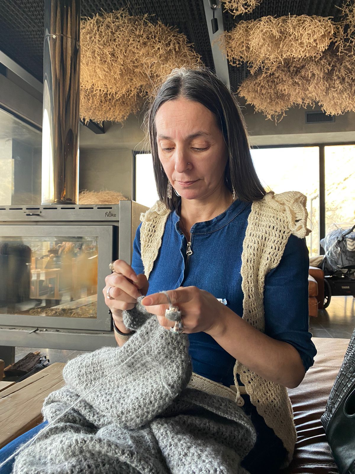 Mujer tejiendo con una aguja, con camisa azul y chaleco color crema. Interior con chimenea y plantas secas colgantes.