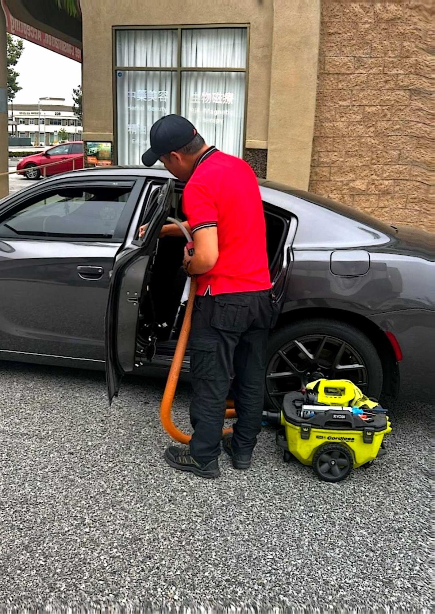 A person in a red shirt and black pants uses a yellow portable vacuum cleaner to clean the interior of a gray car.