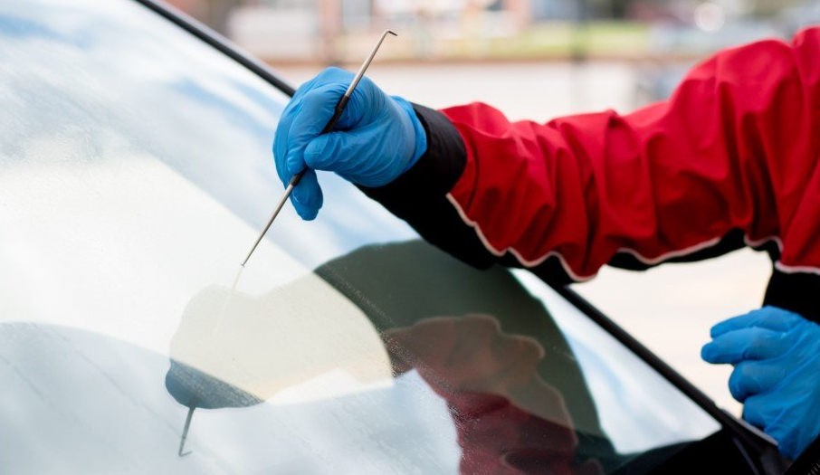 A person in a red jacket and blue gloves uses a tool to examine a car windshield, possibly for damage.
