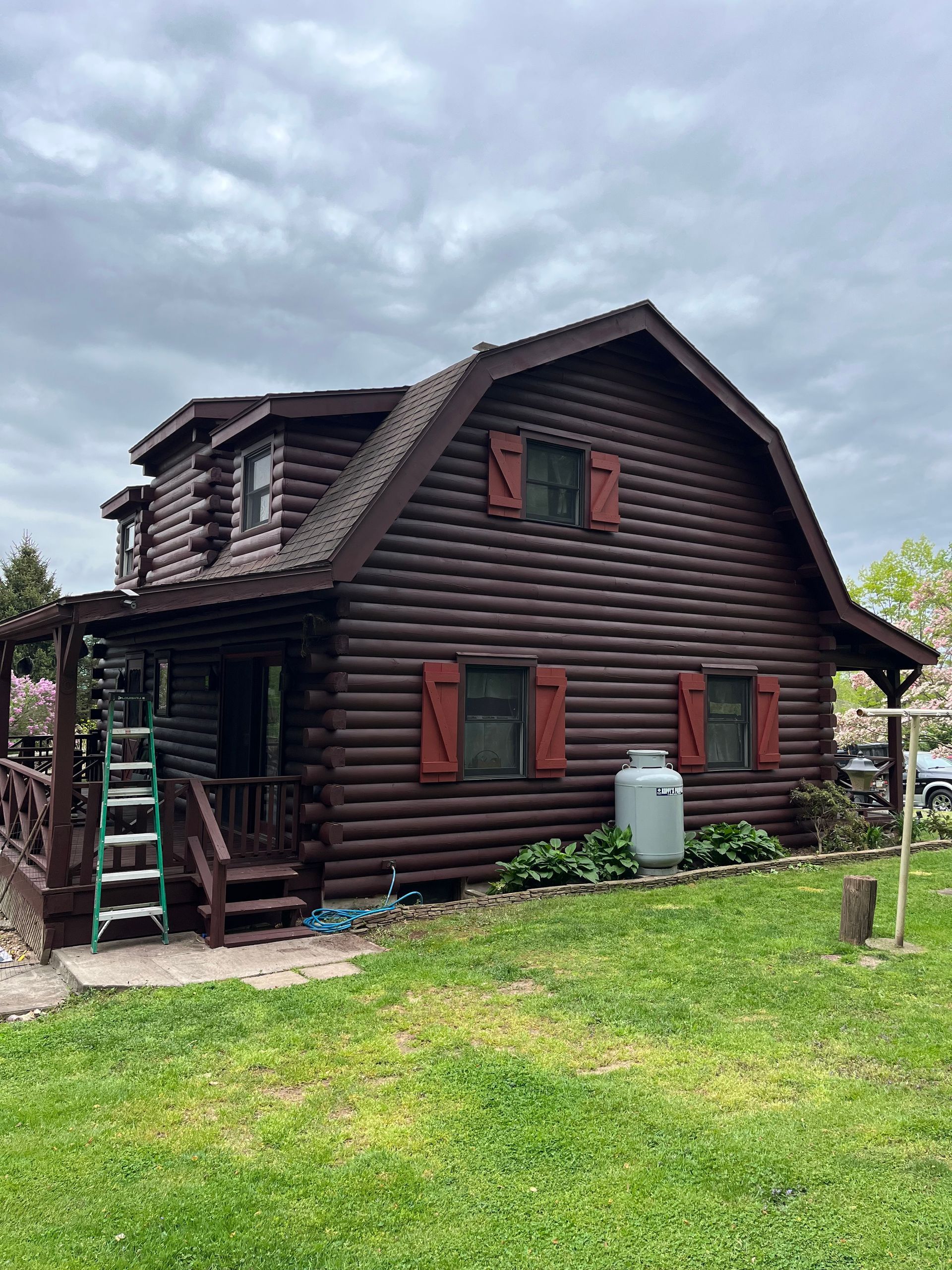 Two-story log cabin with dark brown walls and red shutters. A green yard and cloudy sky are visible.