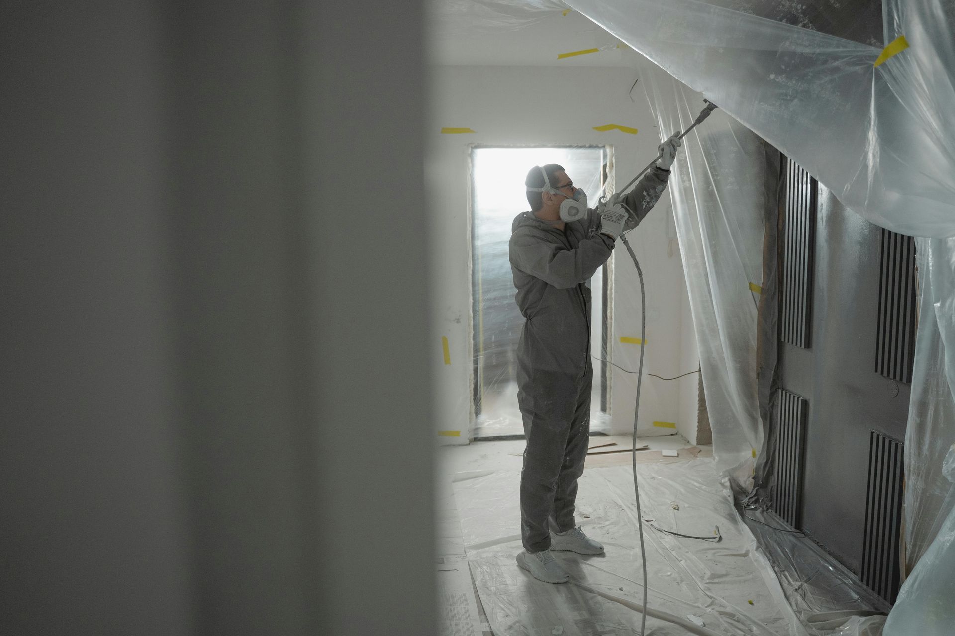 Person in protective suit spraying paint on a wall, covered with plastic sheeting. Indoors, construction site.