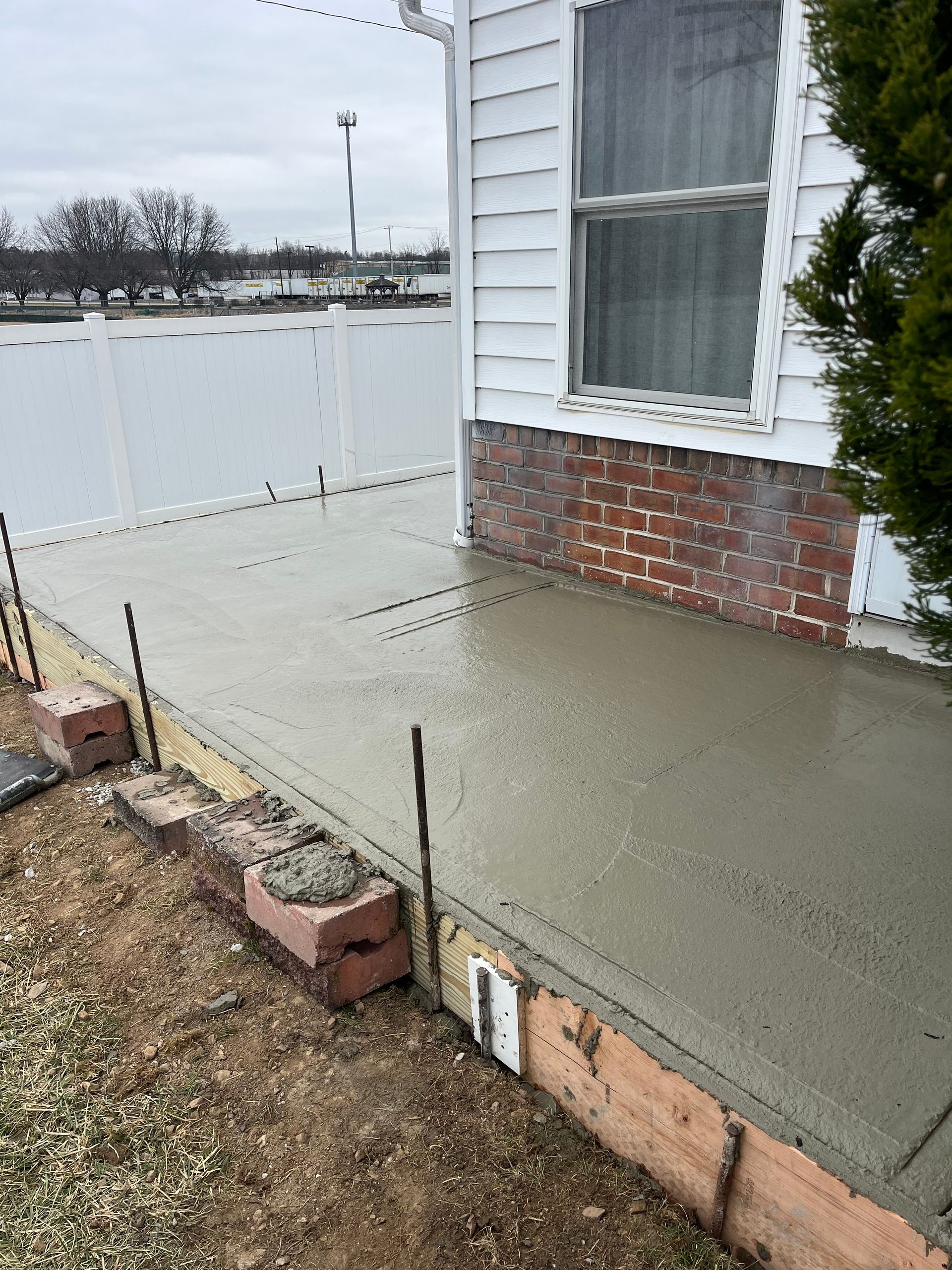 Newly poured concrete patio next to a white house with a window and red brick foundation.
