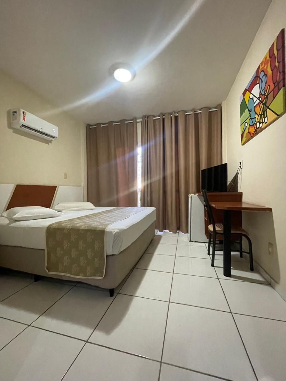 Quarto de hotel pequeno com duas camas, ar condicionado, escrivaninha, cadeira e janela com cortina.