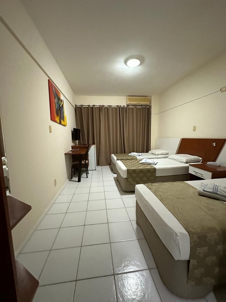 Quarto de hotel pequeno com duas camas, piso de azulejo, janela com cortina e uma escrivaninha encostada na parede.