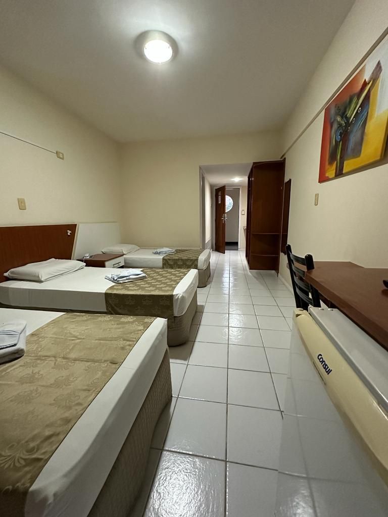 Quarto de hotel pequeno com três camas de solteiro, piso de azulejo e porta aberta para um corredor.