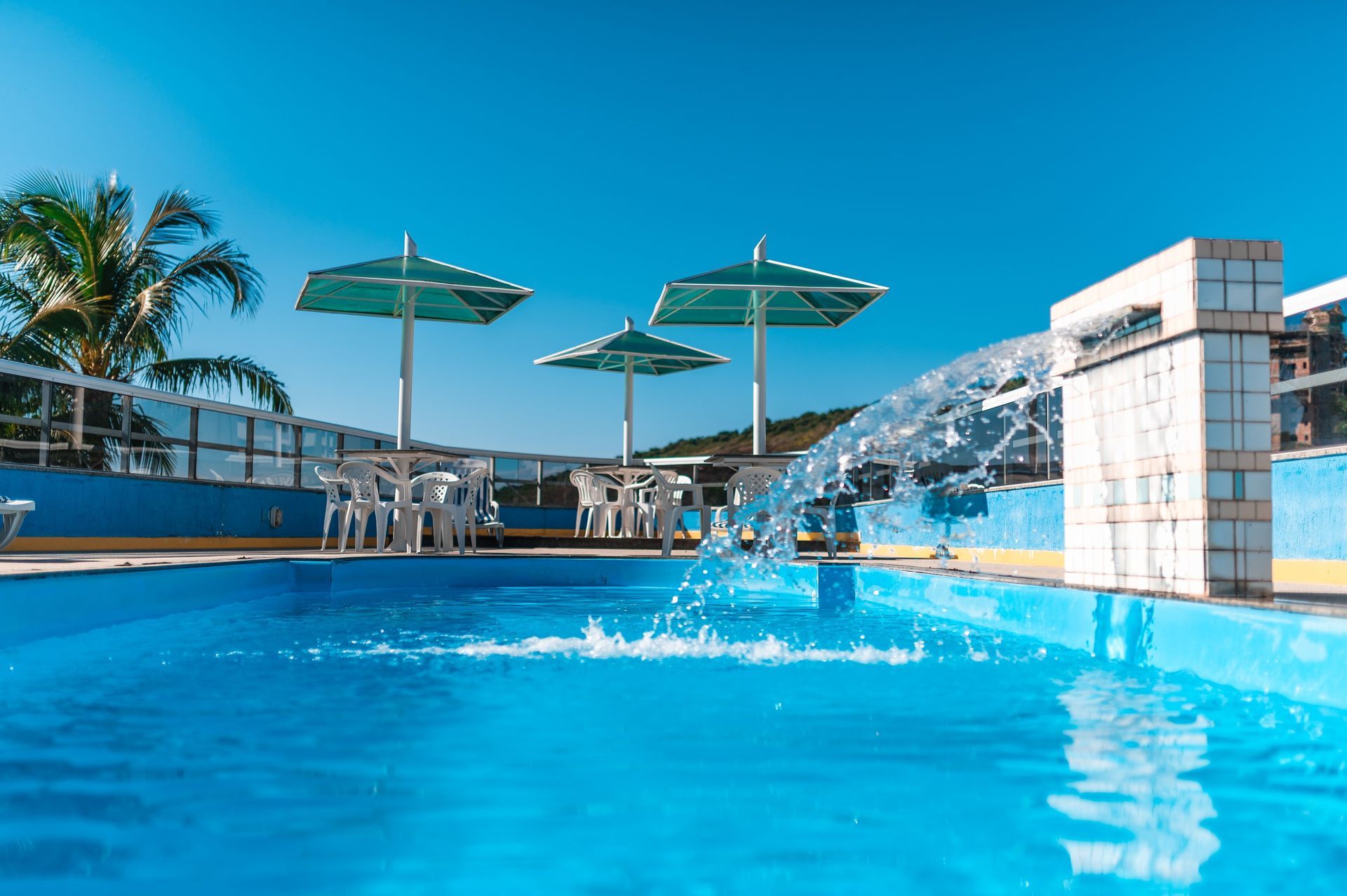 Piscina ao ar livre com água azul cristalina, guarda-sóis, palmeiras e uma montanha nevada ao fundo.