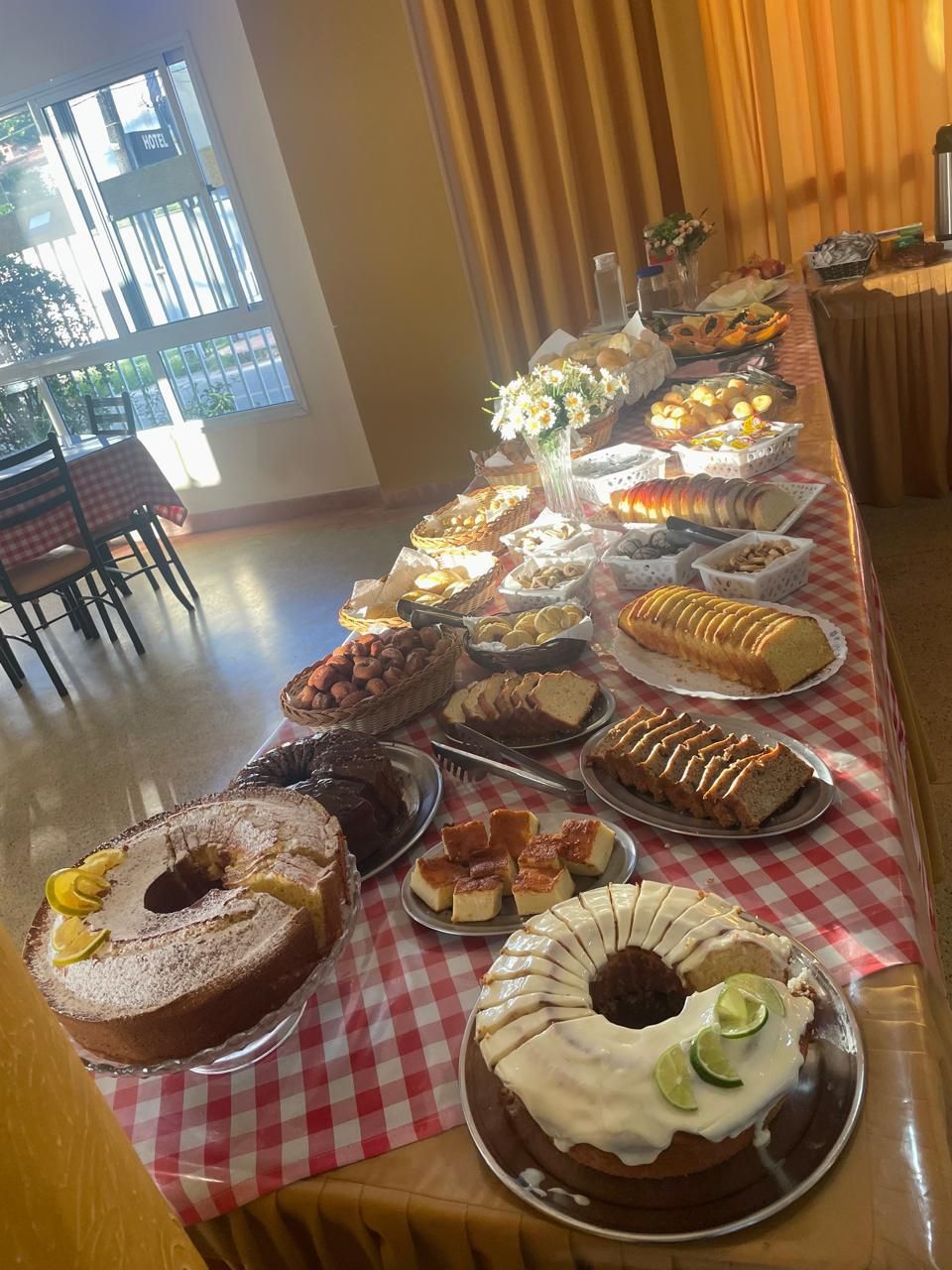 Buffet de sobremesas sobre uma toalha de mesa xadrez com bolos, doces, frutas e flores em um salão de banquetes.